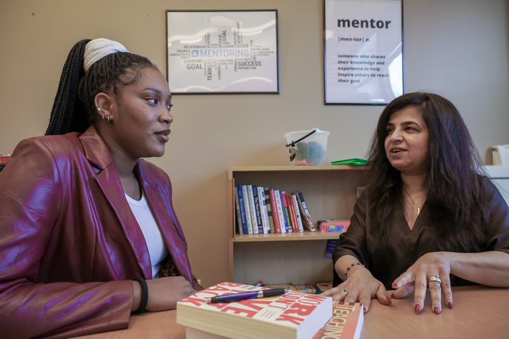 Student and staff member talk at a table with books between them and mentor poster on the wall behind them.