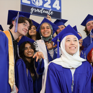 a row of sitting students in graduation regalia with two facing the camera and smiling