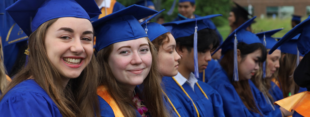 Students sitting in a line wearing blue graduation cap and gowns. Two students on right smiling at the camera.