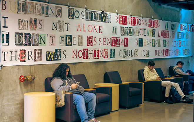 College Policies Students sitting by a wall with a large sign about positive relationships, looking at their phones.