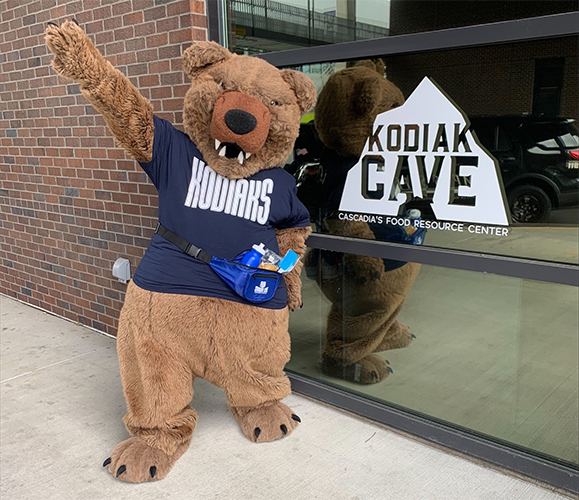 Kodiak Cave: Community Service Cascadia College Kody mascot wearing Cascadia t-shirt and fanny pack waving in front of Kodiak Cave window